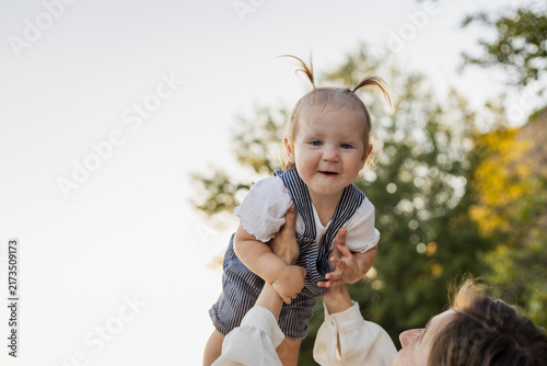 Joyful baby with pigtails held high by parent outdoors in soft natural light