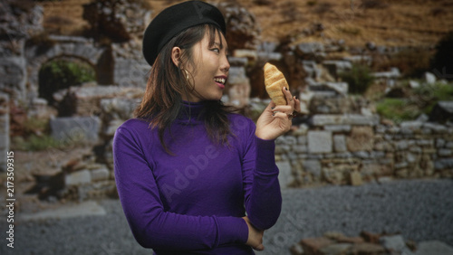 Canvastavla Young asian woman holding a croissant to her ear in a stone ruin building, wearing black beret and purple turtleneck, hand raised near face; playful curiosity