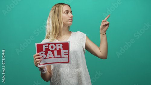 Fotografía Woman blonde holding a for sale sign and pointing finger to the right in studio with teal backdrop, showing a persuasive selling pose; sale negotiation doubt