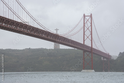 Close view of Lisbon’s 25 de Abril Bridge spanning the Tagus River, with the Cristo Rei monument visible through misty weather beneath a gray overcast sky.