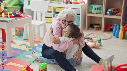 Canvastavla Elderly woman reaches arms to hug little girl on a toy strewn playmat inside a building; affection bonding