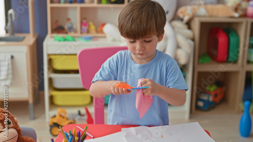 фотографія Boy cutting pink paper with orange scissors using both hands at a play table in a building; concentration creativity