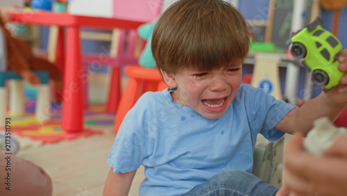 Konstfotografi Boy crying, holds green toy car with hand in a children's playroom building, tears on face and open mouth; distress