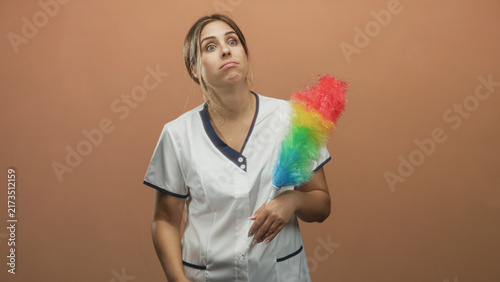 Foto Woman cleaning attendant holding rainbow feather duster and hand to brow showing exhaustion in studio near a plain wall; work fatigue