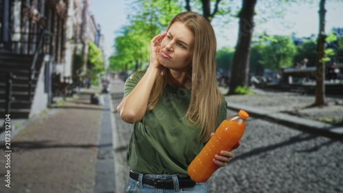 Konstfotografi Woman holding a large orange juice bottle, touching her ear and grimacing while standing on a cobblestone street lined with trees and buildings; discomfort thirst