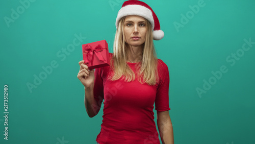 Obraz na płótnie Woman holding a wrapped red gift box with bow and wearing a santa hat and red shirt against a teal studio backdrop; melancholy holiday giving