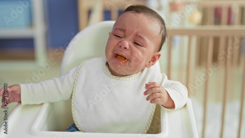Fotografie Baby boy crying and spitting spoonful of puree into mouth with messy bib while seated in highchair in building; feeding frustration