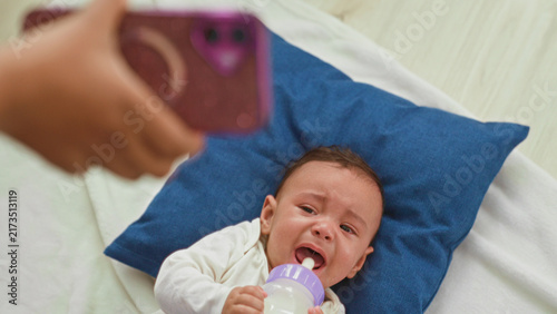 фотографія Hispanic baby boy crying, grasping a milk bottle with mouth open and tears, lying on a blue pillow in a bedroom; distress hunger need fussiness