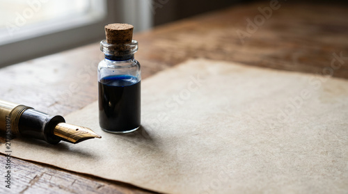 Vintage fountain pen and bottle of blue ink on blank parchment paper over dark wooden table with copy space