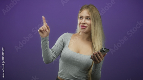 Bild auf Leinwand Young blonde woman pointing index finger while holding smartphone in studio with purple backdrop; playful guidance