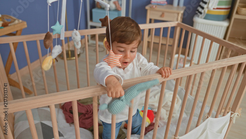 Fotografía Boy stands in wooden crib and reaches for hanging plush mobile while gripping the rail inside a domestic building; gentle curiosity