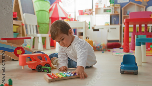 Konstfotografi Boy seated on floor playing a xylophone and pushing a toy bus amid scattered blocks and chairs in a bright playroom; curiosity focus