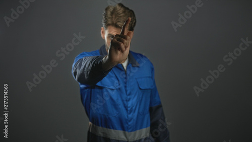 Принти на полотні Man with beard in blue work overalls points index finger forward, inspecting his hand across frames in studio; determination focus resilience