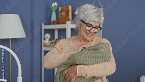 Foto Woman cradling swaddled baby, hand adjusting green blanket and smiling while holding infant near a crib in a building; tenderness bonding