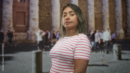 Fotografía Middle age hispanic woman in pink striped shirt looks at camera with slight head tilt and arms relaxed in front of stone columns on crowded street; confidence travel memory heritage