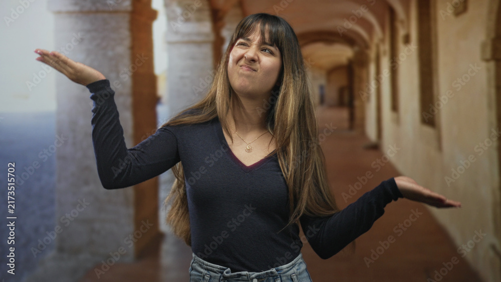 Obraz premium Woman smiling holds out palm and gestures with open hand in a building corridor with arched columns and tiled floor; playful confidence.