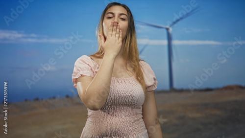 Woman blowing kiss with hand to lips wearing pink dress in studio with wind turbine behind her and open sky; affection calm.