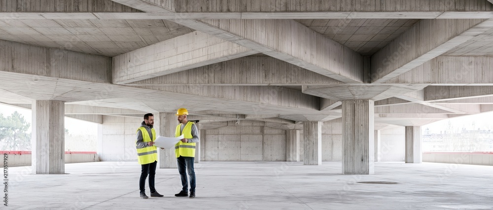 Obraz premium Architect and builder standing at construction site reviewing blueprint under concrete structure