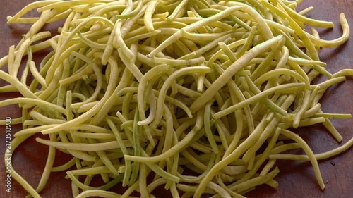 A heap of Golden Teepee yellow wax beans rotating on brown plywood. Fresh, organic, raw, trimmed bean pods pile close-up on a dark wood background, slowly spinning in a tilted view.