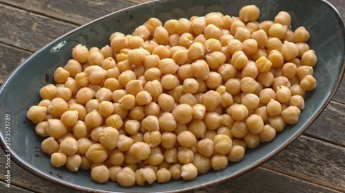 Boiled chickpeas heap rotating on an oval ceramic platter. Fresh organic garbanzo beans, close-up on a rustic dark wood plank background, slowly spinning in a tilted view with natural window light.