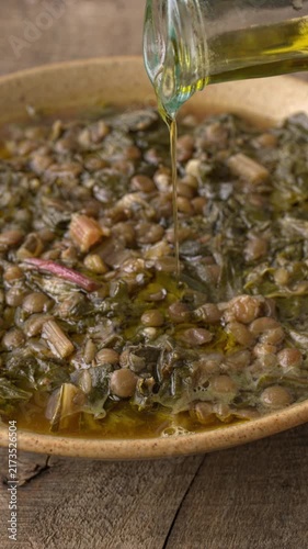 Pouring an olive oil drizzle from a bottle onto a thick, homemade, traditional Mediterranean soup. Greek Faki me Seskoula lentil stew with Swiss chard on rustic wood. Vertical video.