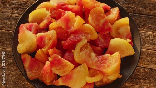 Old German tomato wedges rotating on a black plate. Sliced organic orange-red heirloom tomato chunks on a rustic dark wooden background in warm late-afternoon golden light.