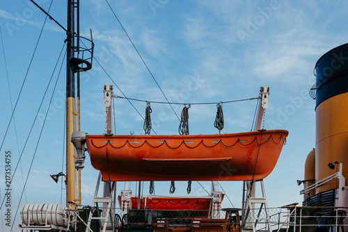 Foto An orange lifeboat hanging on davits on a ship deck near a large yellow funnel under a blue sky with soft clouds