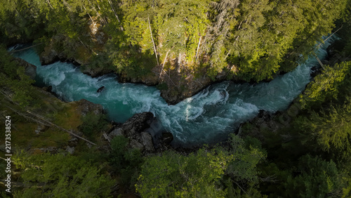Obraz na płótnie A straight top down aerial wide shot overlooks a tiny wooden footbridge spanning across the roaring white water rapids of the turquoise Cheakamus River inside a dense pine forest