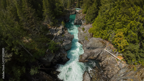 An aerial wide shot captures the fast flowing turquoise water of the Cheakamus River rushing over white water rapids inside a deep forest canyon on a bright day.