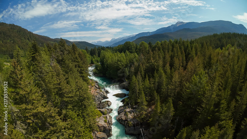 Konstfotografi An aerial wide shot looks across a lush evergreen pine forest valley to reveal the rushing cascades of Cheakamus River Waterfalls beneath rolling mountain peaks under a cloudy sky