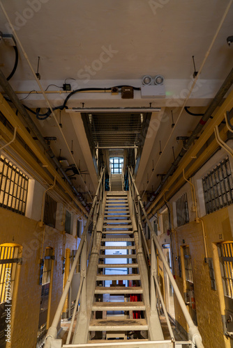 Fotografie Direct upward view of a long metal staircase connecting multi-story cellblock galleries with yellow walls, barred doors, and institutional architecture