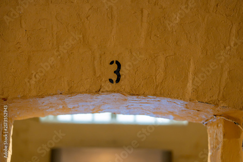Obraz na płótnie A stark black stenciled number three on a heavily painted, textured yellow stone arch over a doorway, with a blurred window in the background