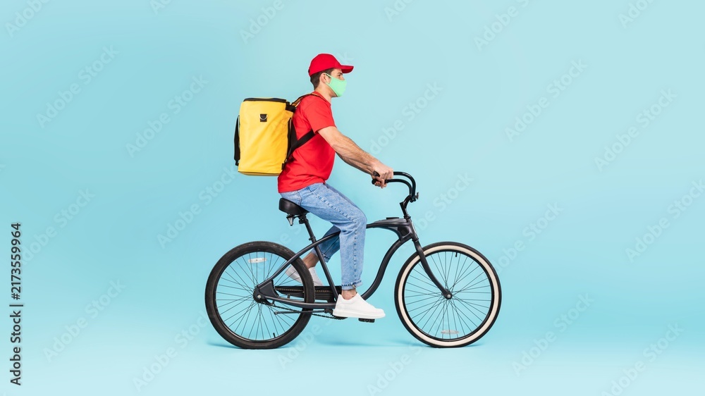 Fototapeta premium Focused Delivery Man Riding Bicycle With Yellow Delivery Bag, Wearing Face Mask, In Urban Environment. Promoting Health And Safety While Providing Delivery Services Outdoors.