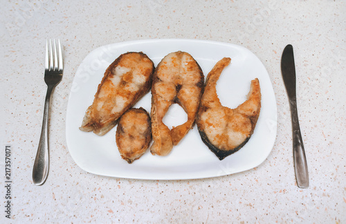 Pieces of fish fried in vegetable oil lie on a white plate, near a fork and knife. Concept for breakfast, lunch, or dinner.