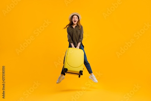Foto Excited Young Caucasian Female Smiling While Jumping With Yellow Suitcase On Bright Yellow Background, Signifying Joyful Travel Experience In A Fun, Vibrant Setting