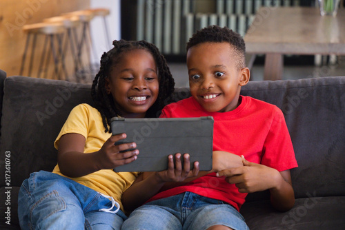 Quadro su tela African American children sitting on dark gray sofa sharing tablet with protecti