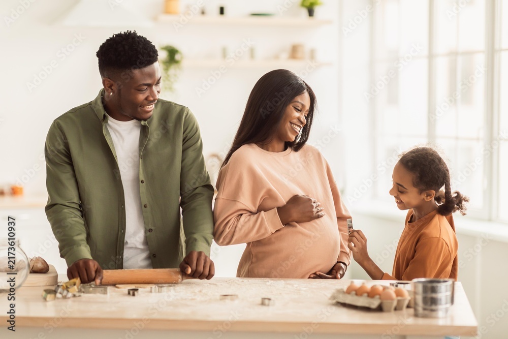 Fototapeta premium Smiling African American Couple With Pregnant Woman And Young Girl Helping In Kitchen, Preparing Food Together, Enjoying Family Time During Cooking In Modern Home. Family Concept.