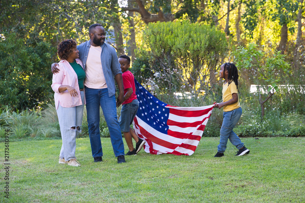 Obraz premium African American family of four walking on grassy lawn in parklike backyard carrying American flag