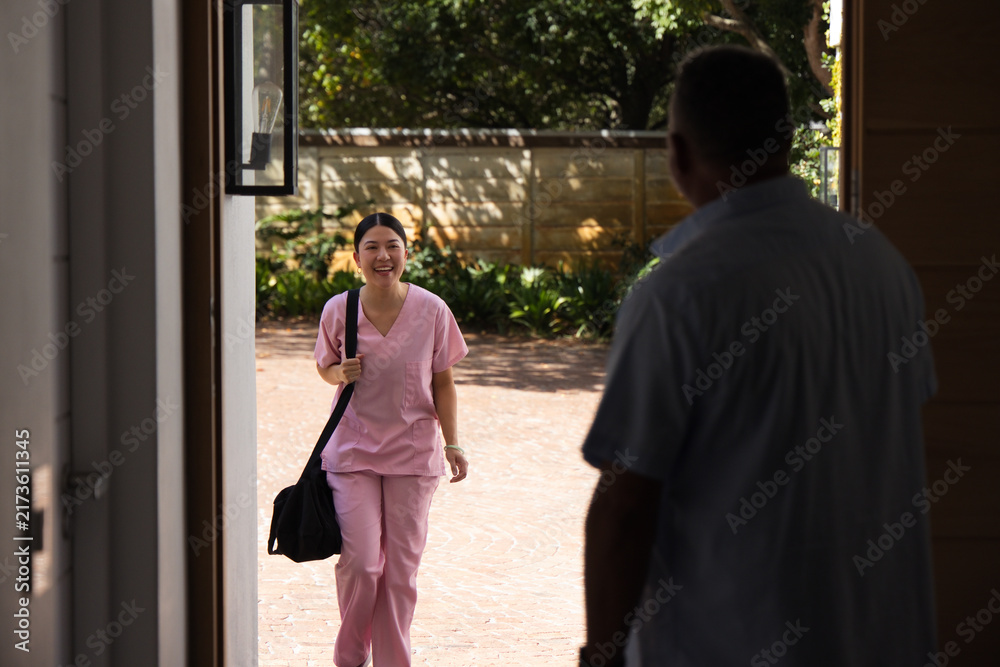 Naklejka premium Asian nurse in pink scrubs walking brick drive to home door carrying black duffel greeting senior