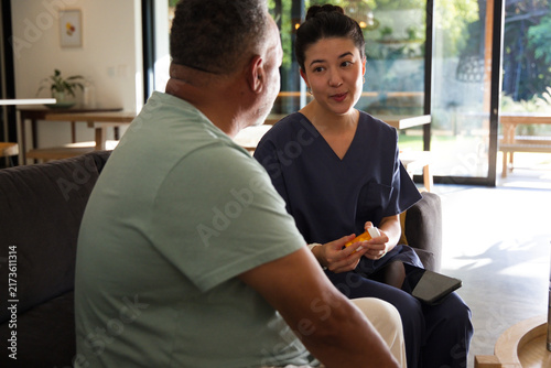 Asian nurse in navy scrubs holding pill-bottle and tablet speaking to client on sofa, copy space