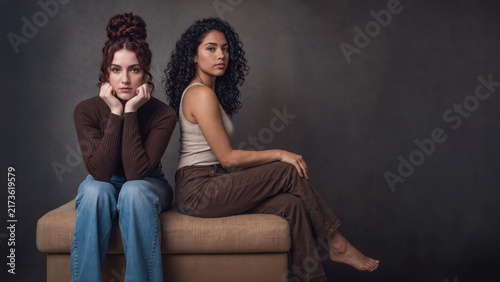 Two fashion models pose on an ottoman in a moody studio portrait, one facing forward with her chin in her hands and the other seated sideways with crossed legs.