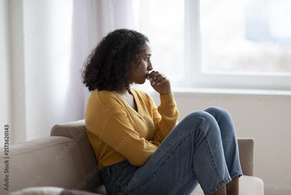 Obraz premium Worried Expression. Black Female Adult, Young Age, Sitting On Couch In Living Room At Home, Staring Out Of Window, Deep In Thought, Reflecting. Concept Of Mental Wellness And Emotional Balance.