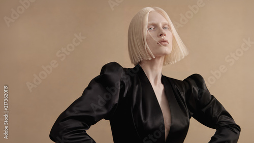 A pale-skinned blonde fashion model with a sleek bob poses confidently in a dramatic black satin dress against a warm neutral studio setting.