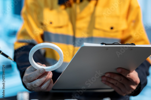Close-up of a utility worker in a yellow safety jacket holding a roll of white tape and a clipboard during an outdoor equipment inspection.
