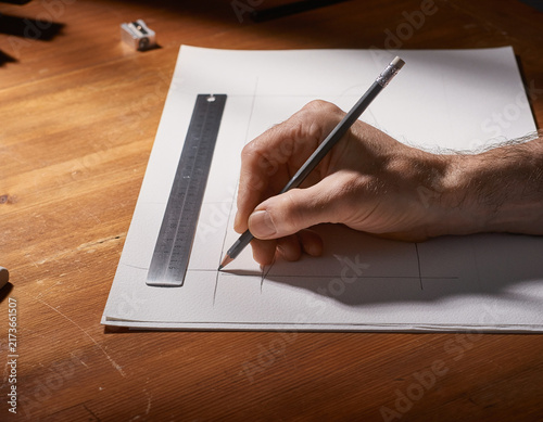 木製デスクの紙の上で鉛筆と定規を使って直線や図面を描く人の手元と製図作業のイメージ