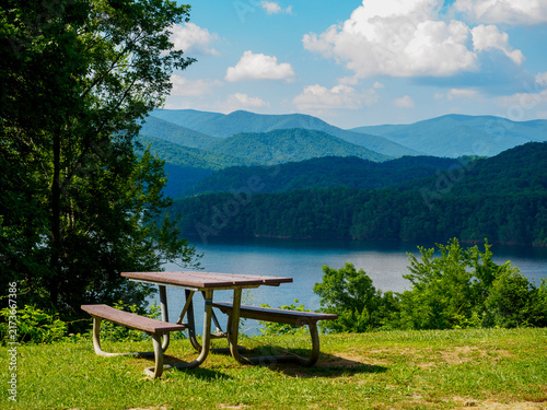 Fotografía A wooden picnic table sits on a grassy overlook over a breathtaking view of a  blue lake surrounded by  forest and mountain ridges