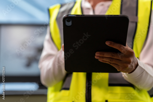 Close-up of a construction worker in a yellow safety vest holding a tablet at an industrial site, showing digital inspection and project management.