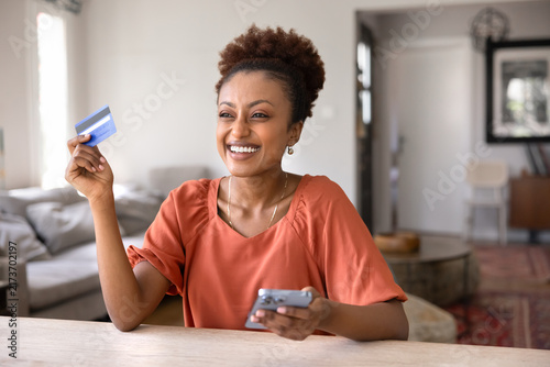 Joyful young African woman holding credit card and smartphone  smiling while making online purchase from home. Mobile banking usage  convenient secure payments  financial success and digital lifestyle
