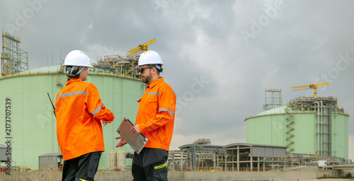 Teamwork technician discussing petroleum engineering in oil refinery project collaboration at fuel industrial plant Male and female engineers holding laptop talking near petrol storage tanks