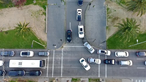 top-down drone footage showing cars and a bus navigating a busy urban intersection surrounded by sidewalks, palm trees and green areas, illustrating traffic flow, commuting and road infrastructure.